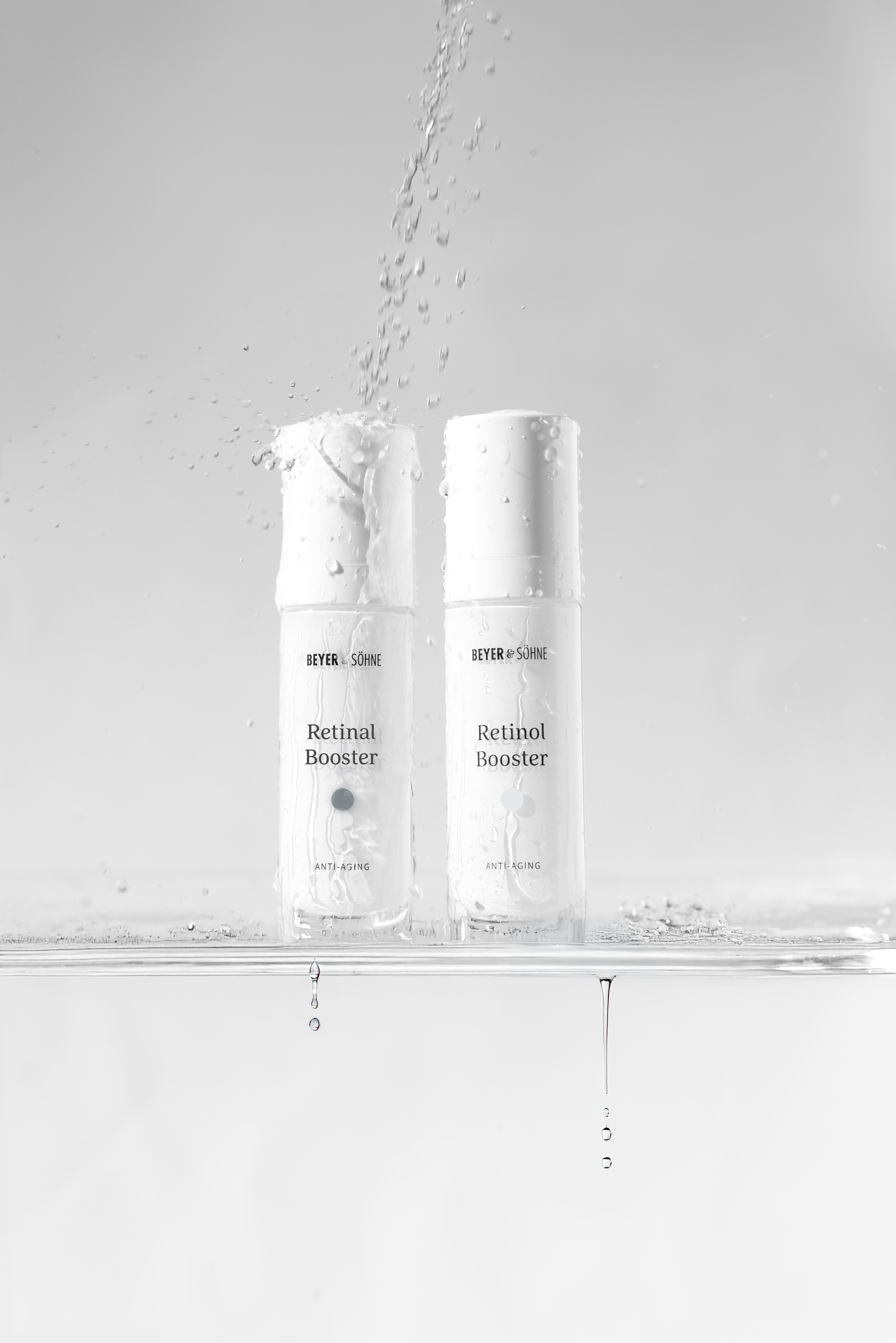 Der Beyer & Söhne Retinal und Retinol Booster stehen nebeneinander auf einer Glasfläche, während Wasser darüber spritzt und Tropfen herablaufen und eine frische, pflegende Wirkung darstellen